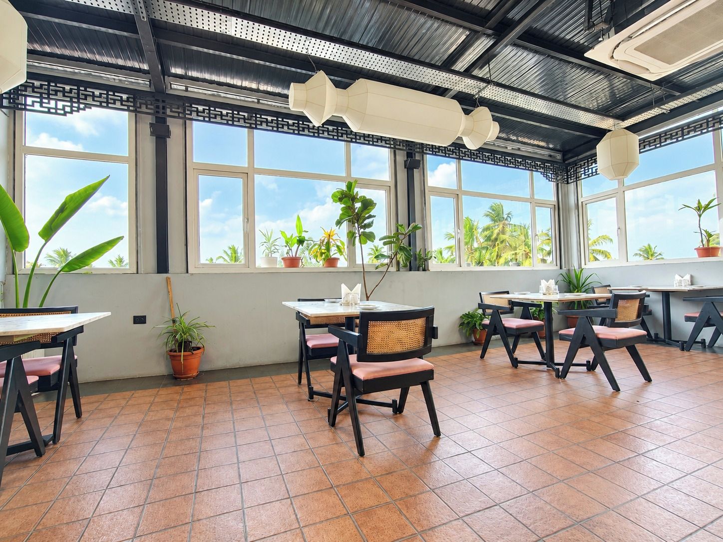 A restaurant with multiple tables and chairs, large windows with views of palm trees, and potted plants.