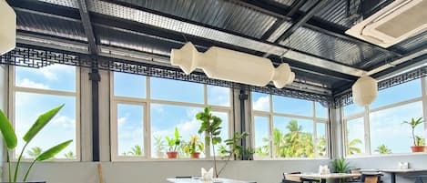 Un restaurante con varias mesas y sillas, ventanas amplias con vistas a palmeras y plantas en macetas.