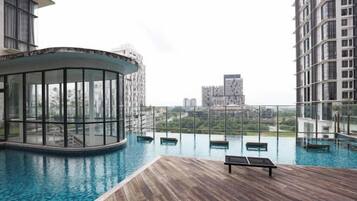 Una piscina infinita en la azotea con terraza de madera, zona de estar cubierta con vidrio y vistas de edificios circundantes.