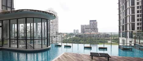 A rooftop infinity pool with a wooden deck, a glass-enclosed lounge area, and a view of surrounding buildings.