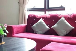 Een rode bank met twee witte kussens, een ronde tafel en een raam met gordijnen.