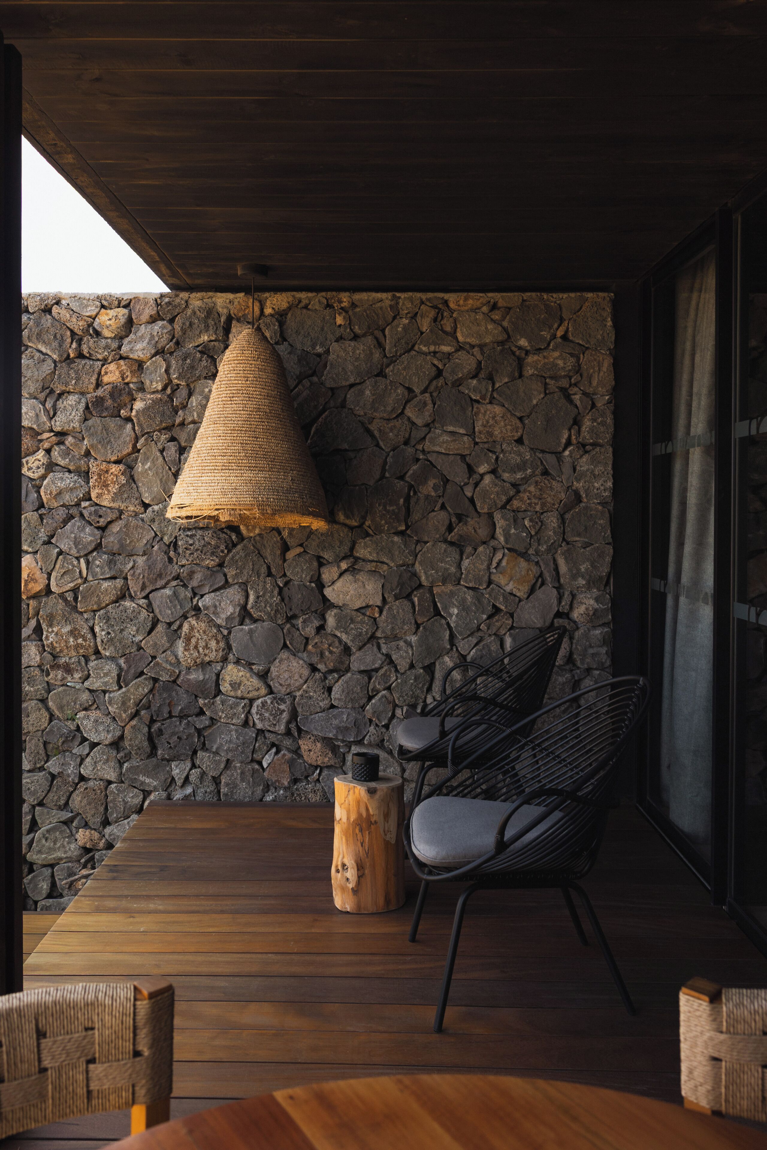 A wooden deck with a stone wall, a woven pendant light, a black chair, and a wooden stool.