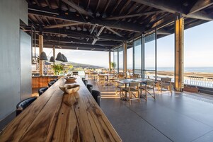Uma ampla sala de jantar com uma longa mesa de madeira e cadeiras, grandes janelas com vista para o mar e um ventilador de teto.