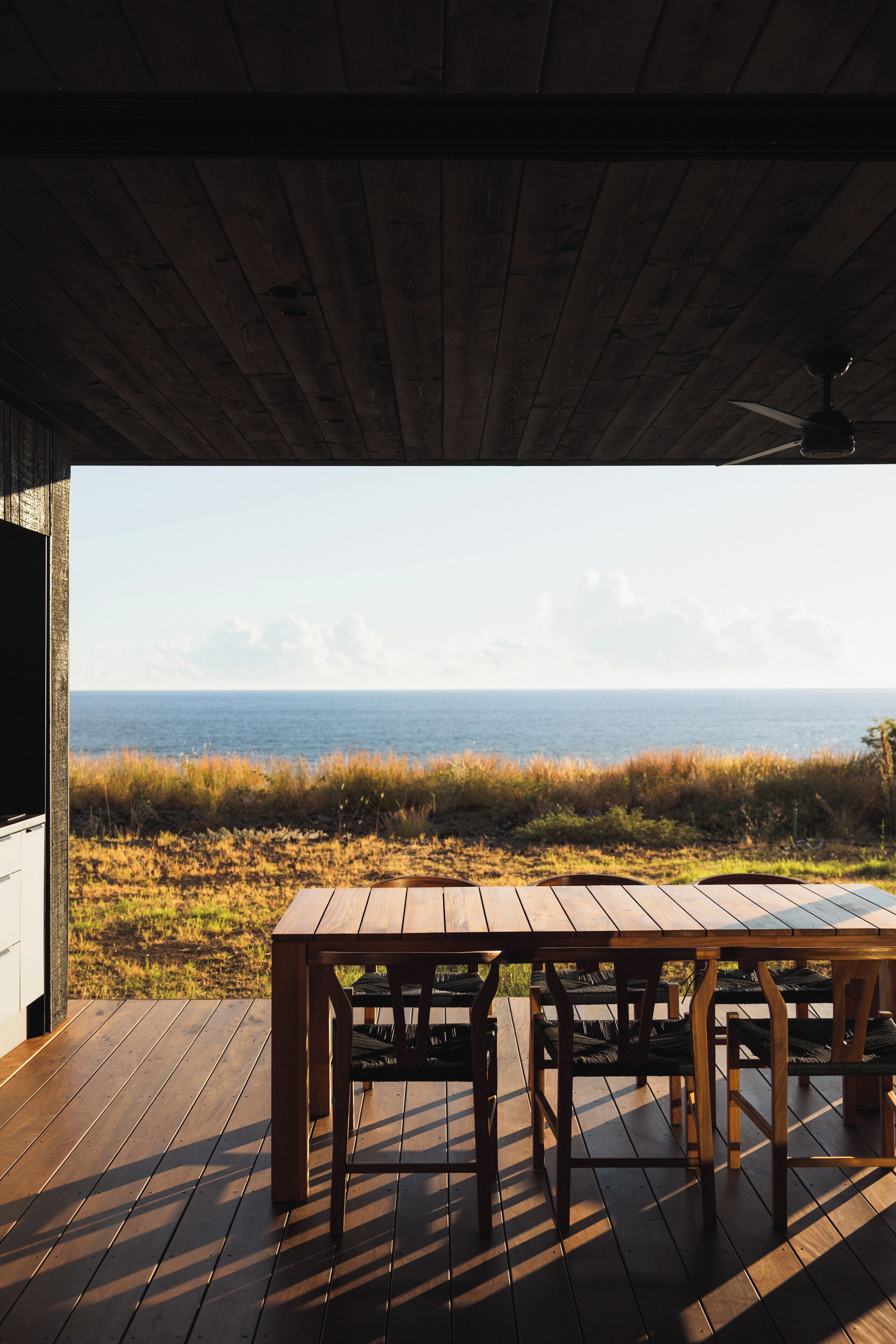 A wooden deck with a table and chairs, overlooking a grassy area and the ocean.