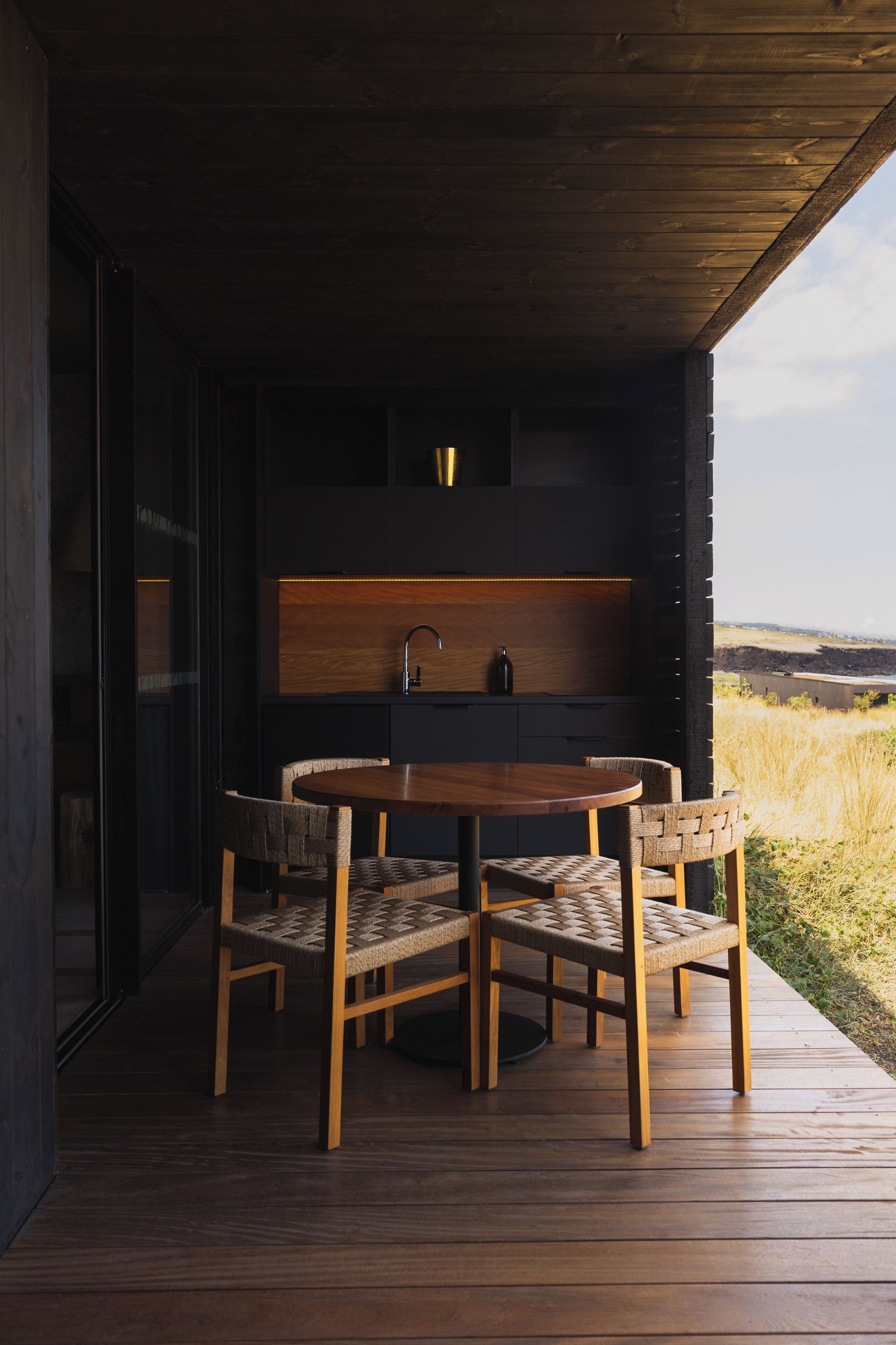 Eine Holzterrasse mit einem runden Tisch und Stühlen, eine integrierte Küche mit Spüle und Schränken sowie ein Blick auf die Landschaft.