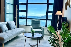 A modern living room with a sofa, armchair, round coffee table, and large windows offering a view of the sea. - L Homestay Jesselton Quay (Near Gaya Street, Near Pier) (Kota Kinabalu)
