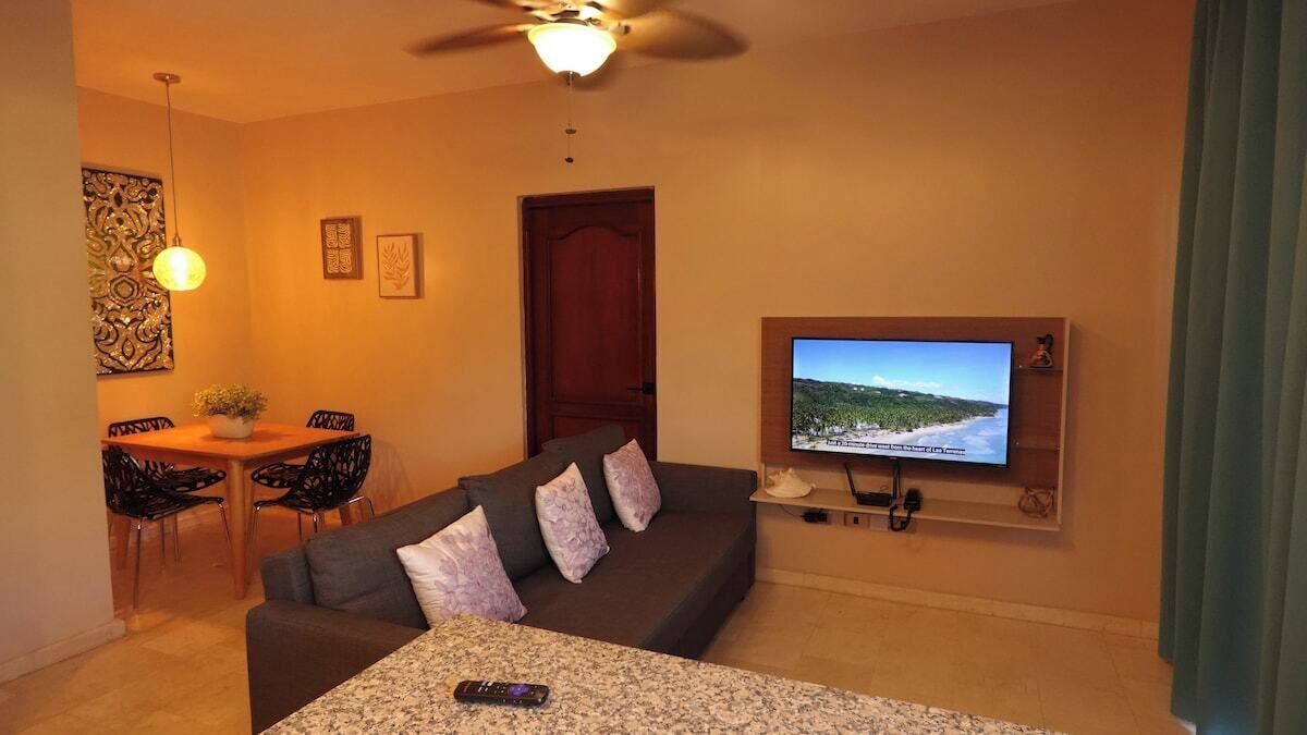 Un salon avec un canapé gris foncé, un téléviseur fixé au mur, un ventilateur de plafond et un coin repas comprenant une table et des chaises.