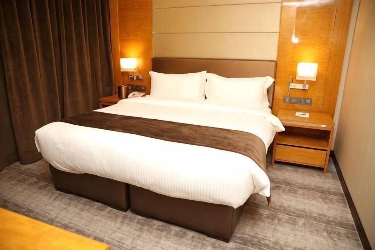 A hotel room with a large bed, two bedside tables, a nightstand, and a wall-mounted lamp.