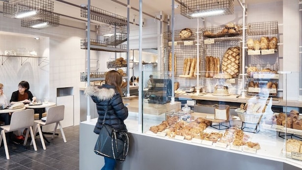 Une boulangerie où l’on peut admirer une variété de pains et de pâtisseries présentés dans des vitrines, un tableau affichant les prix et un espace avec des tables et des chaises où l’on peut s’asseoir.