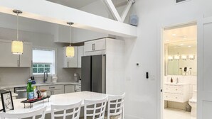Een moderne keuken met witte kasten, een centraal eiland met barstoelen, hanglampen en uitzicht op de aangrenzende badkamer.