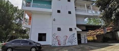 Un edificio de dos pisos con un balcón en el nivel superior, un coche estacionado en la parte delantera y una pared decorativa con un diseño similar a alas.