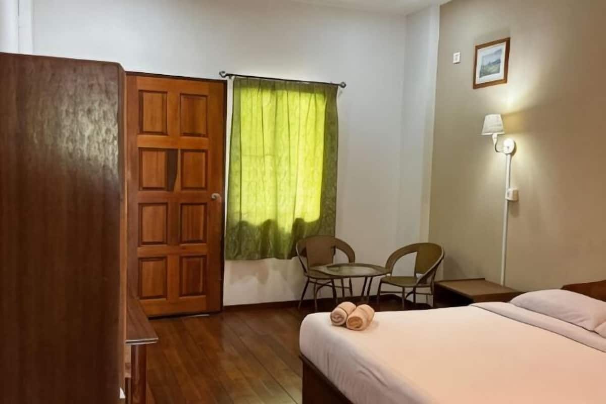 Ein Hotelzimmer mit einem Holzschrank, einem Bett mit weißer Bettdecke, zwei Stühlen, einem kleinen Tisch, einem grünen Vorhang und einer Wandlampe.