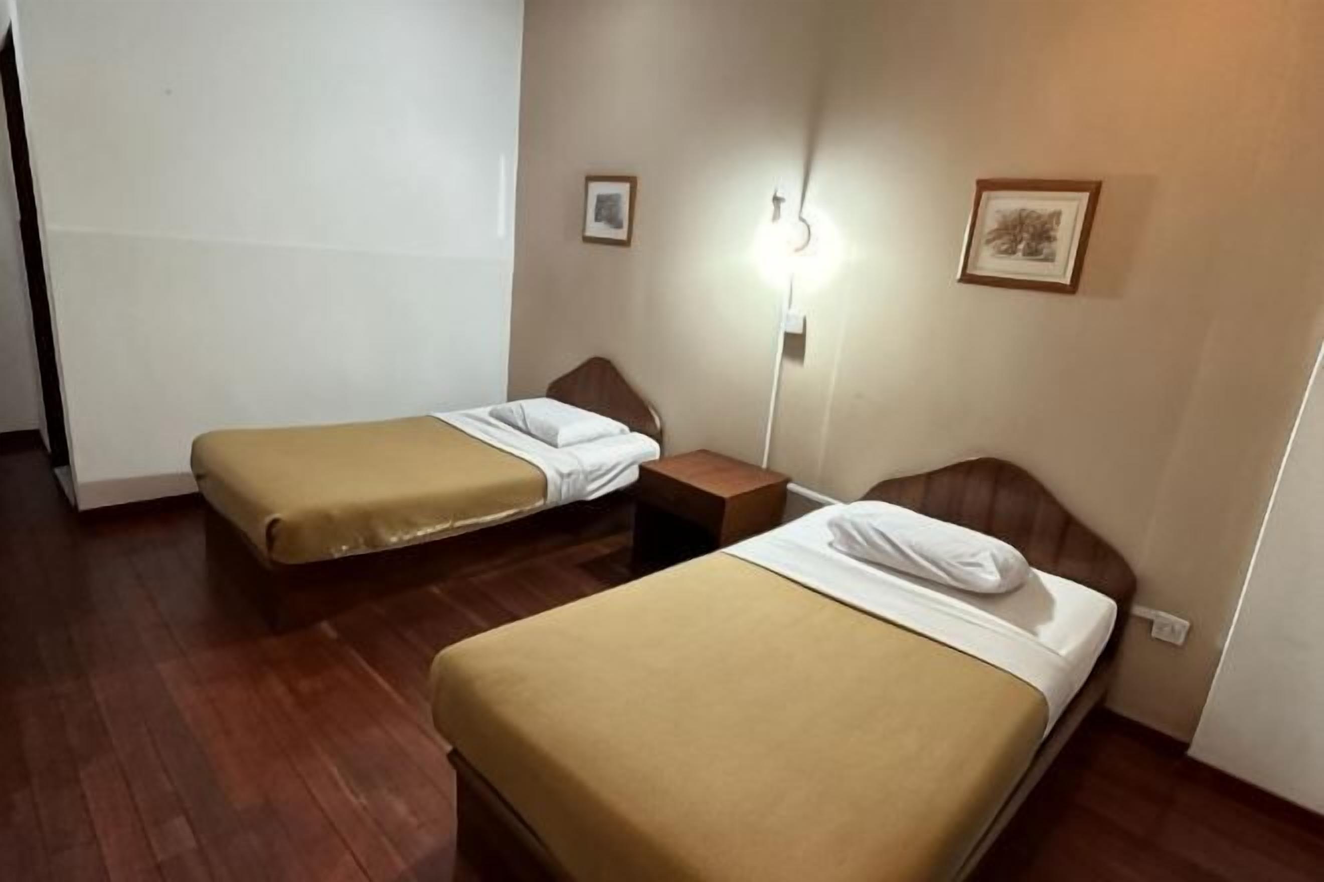 Una habitación con dos camas individuales, una mesita de noche, una lámpara de pared y dos cuadros enmarcados.