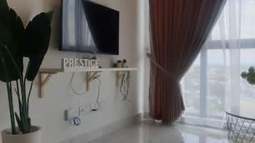 A modern living room with a flat-screen TV mounted on the wall, a potted plant, a shelf with decorative items, and a large window with curtains.
