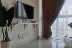 Uma sala de estar moderna com televisão de tela plana na parede, uma planta em vaso, uma estante com objetos decorativos e uma janela grande com cortinas.