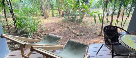 A patio with two wooden lounge chairs and a wicker table, surrounded by lush greenery and a small garden area.