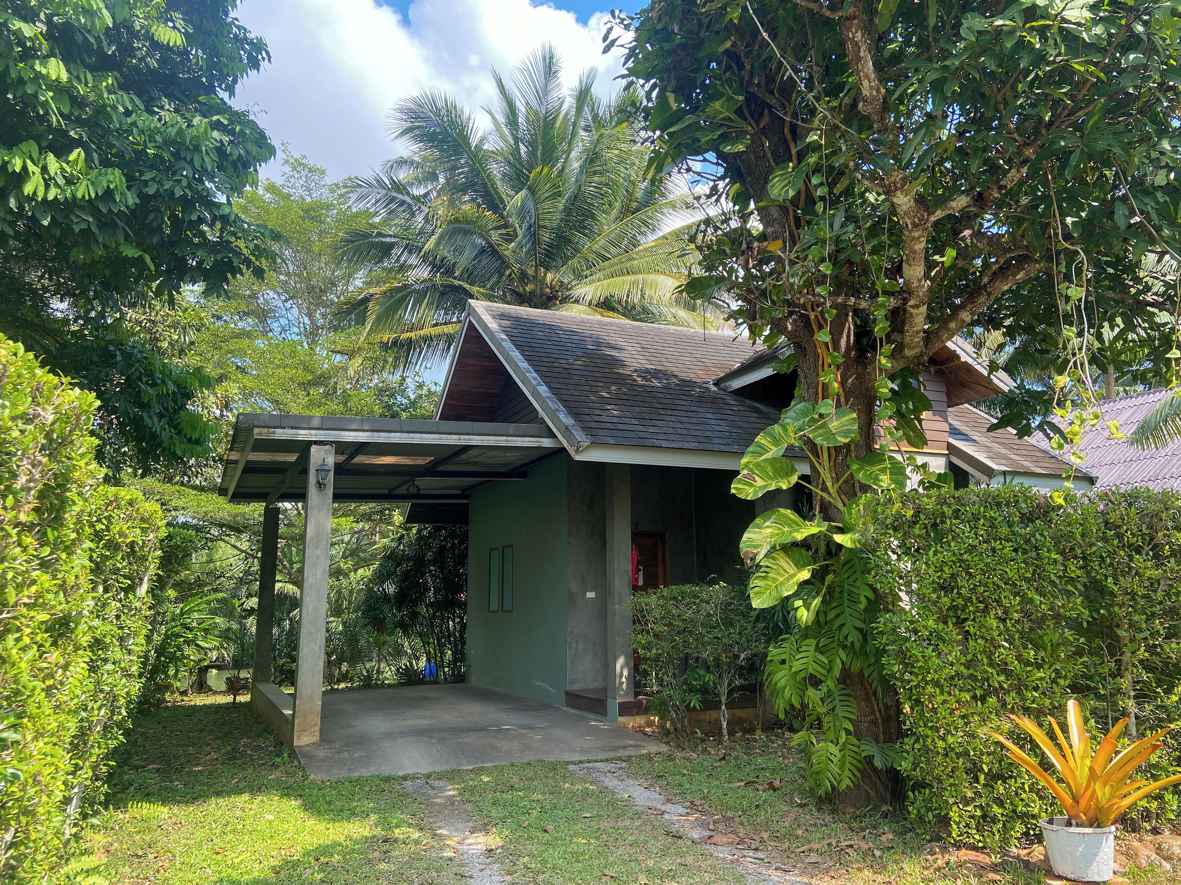 A small, single-story building with a sloped roof, surrounded by lush greenery and trees.