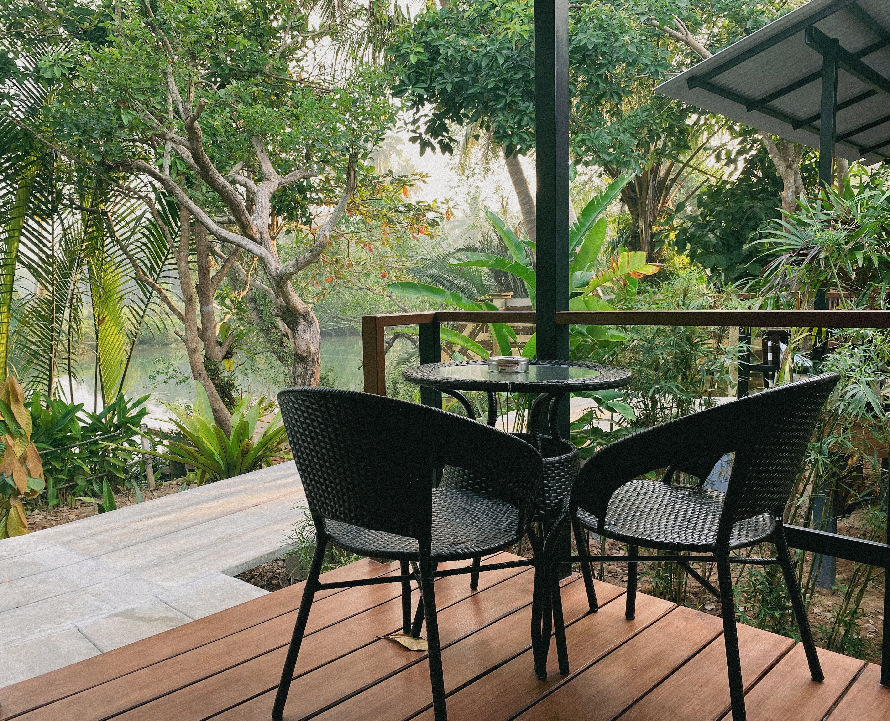 An outdoor patio with a table and chairs, surrounded by lush greenery and a view of a body of water.