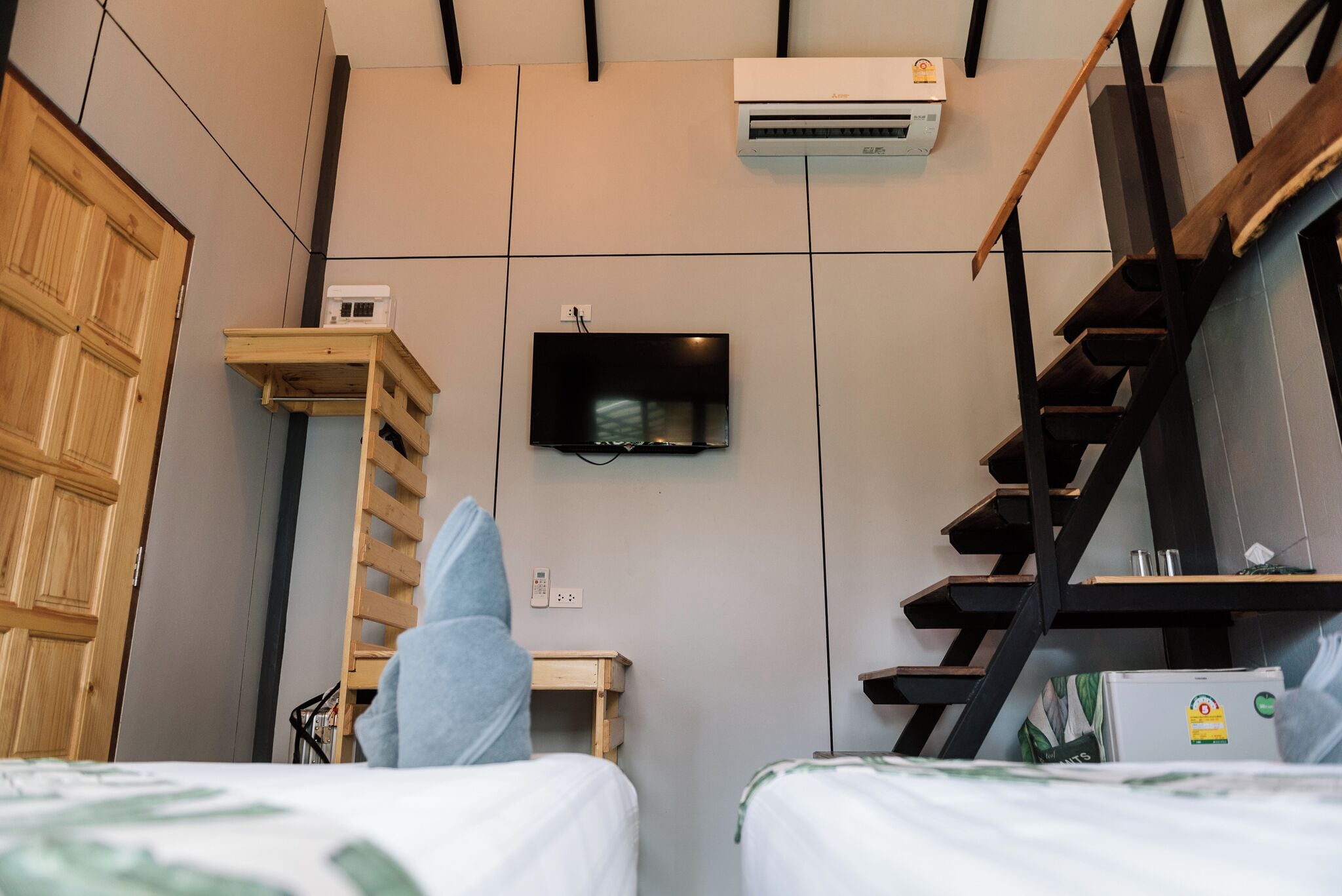Un dormitorio con dos camas, un armario de madera, un televisor montado en la pared, una escalera y un equipo de aire acondicionado.