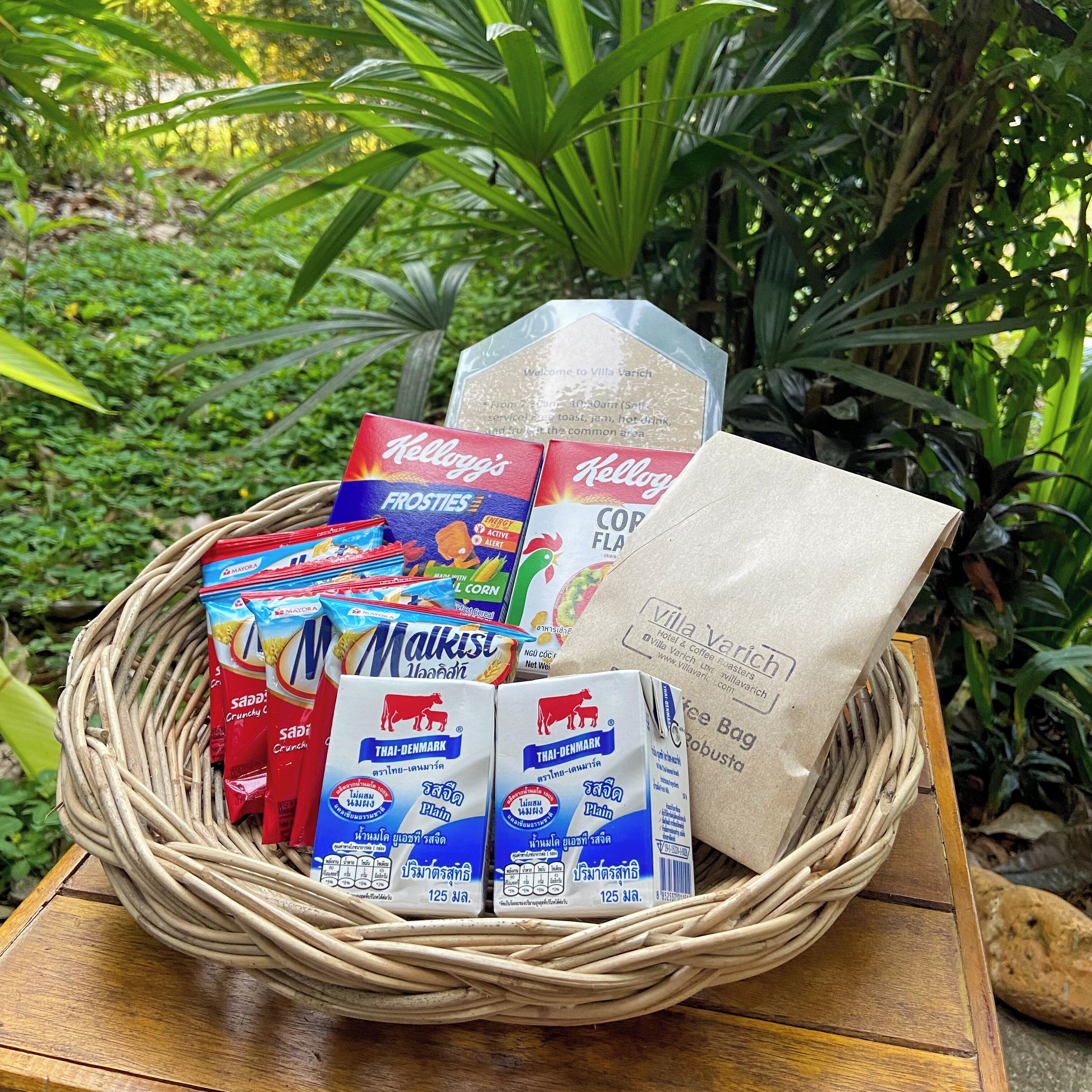 Una cesta de mimbre con diversos alimentos, incluyendo el cereal Kellogg's Frosties, leche Thai-Denmark y una bolsa de café.