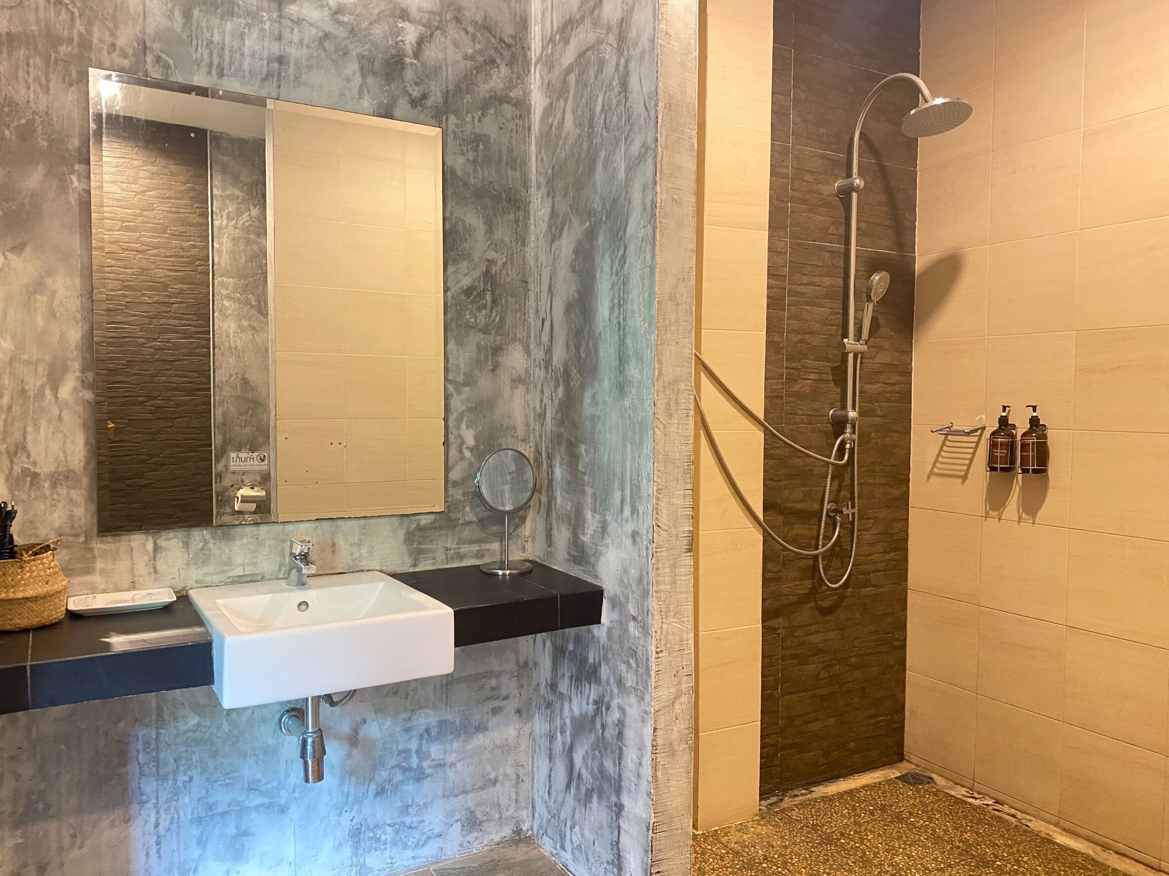 A bathroom with a white sink, a mirror, a handheld mirror, a shower with a handheld showerhead, and two bottles of shampoo.