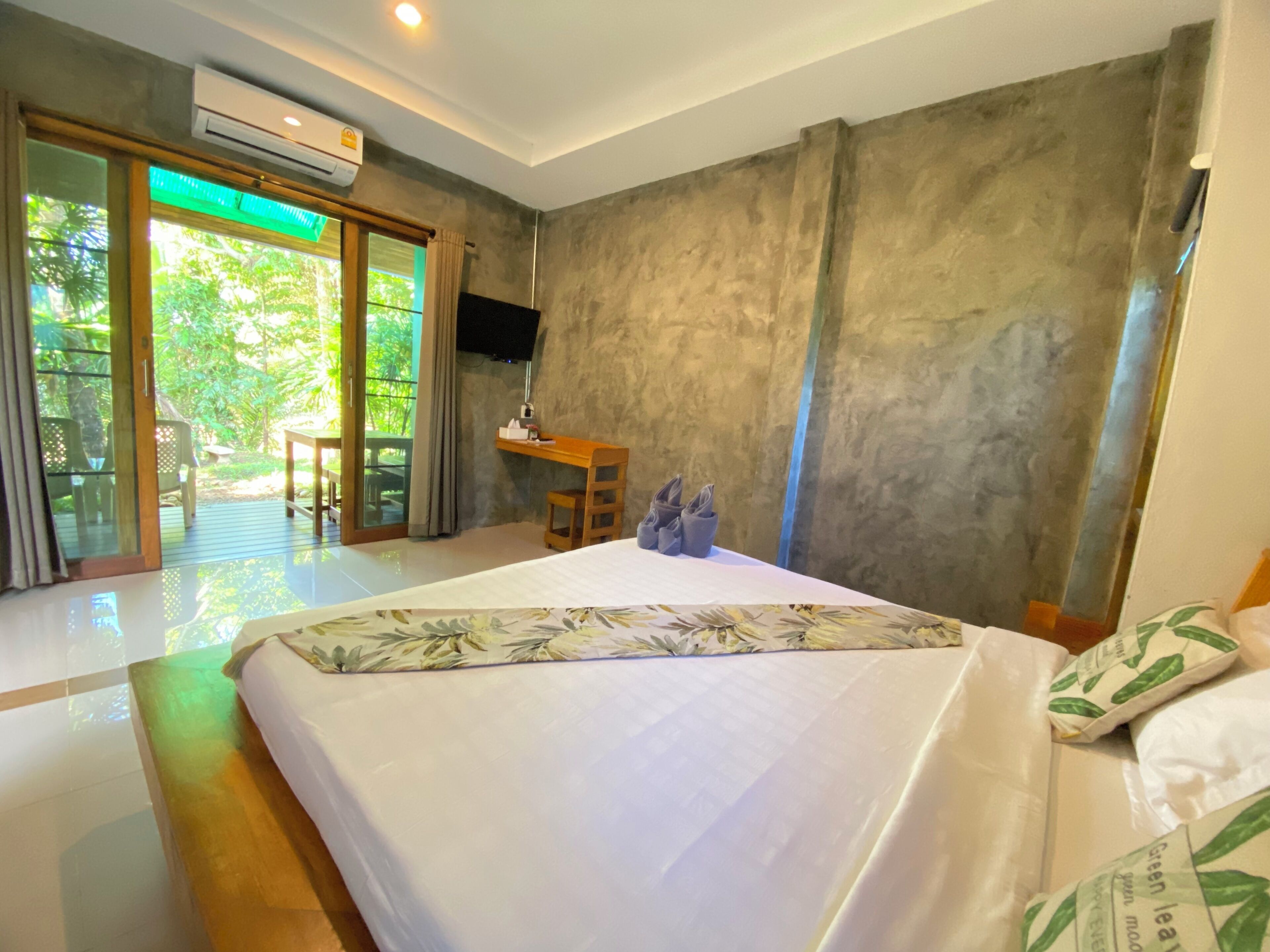 A hotel room with a bed, a small wooden table, a TV mounted on the wall, and a sliding glass door leading to an outdoor area.