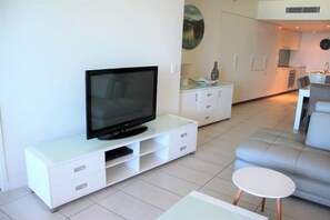 Een moderne woonkamer met een flatscreen televisie op een wit kastje, een grijze bank en een kleine witte tafel.