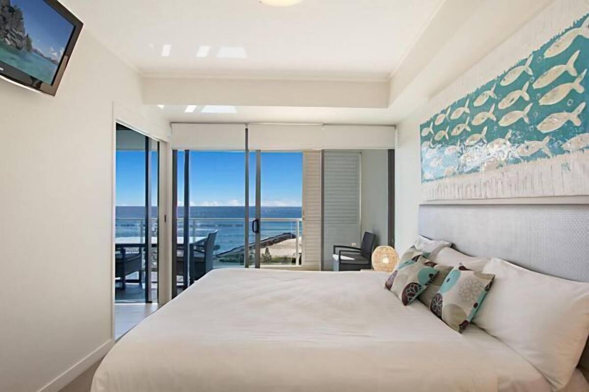 A bedroom with a large bed, a view of the ocean, a painting of fish, and a chair by the window.