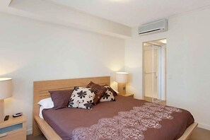 A bedroom with a wooden bed, two bedside lamps, a nightstand, and a door leading to another room. - Blue C Coolangatta (Coolangatta)