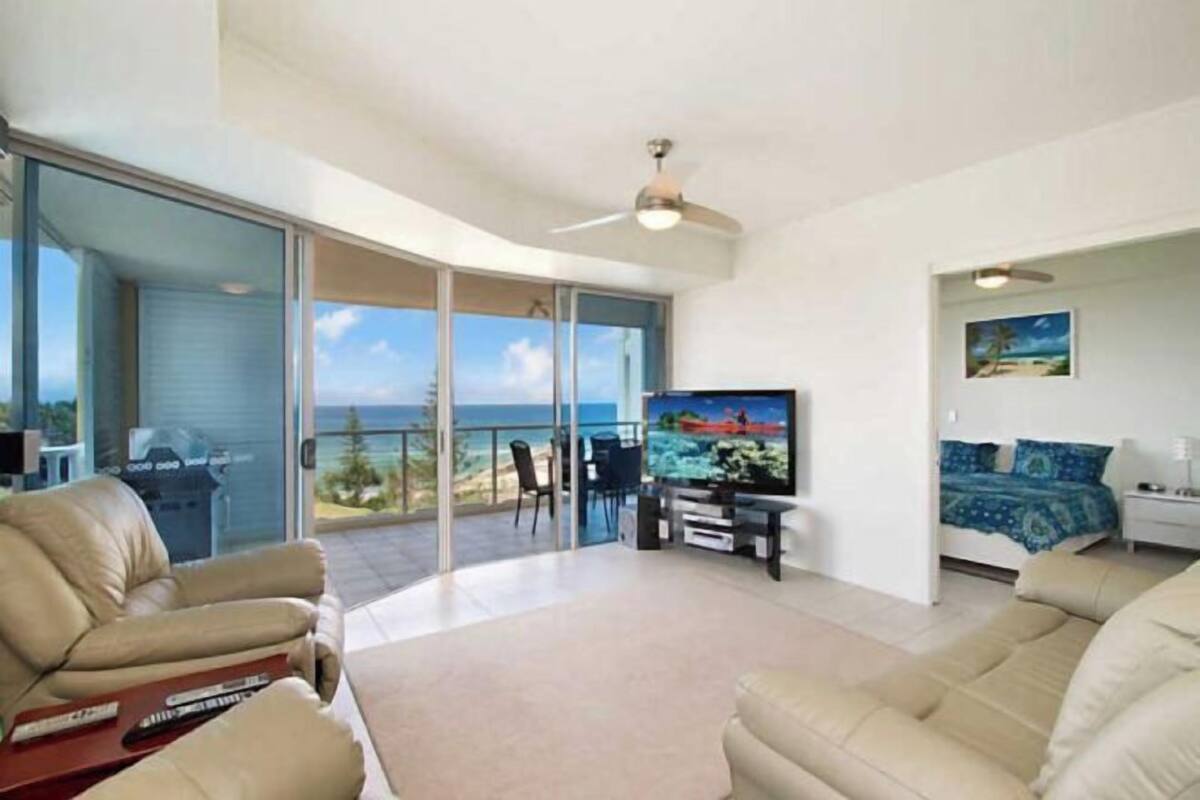 A modern living room with a large TV, comfortable seating, a balcony with a view, and a ceiling fan.