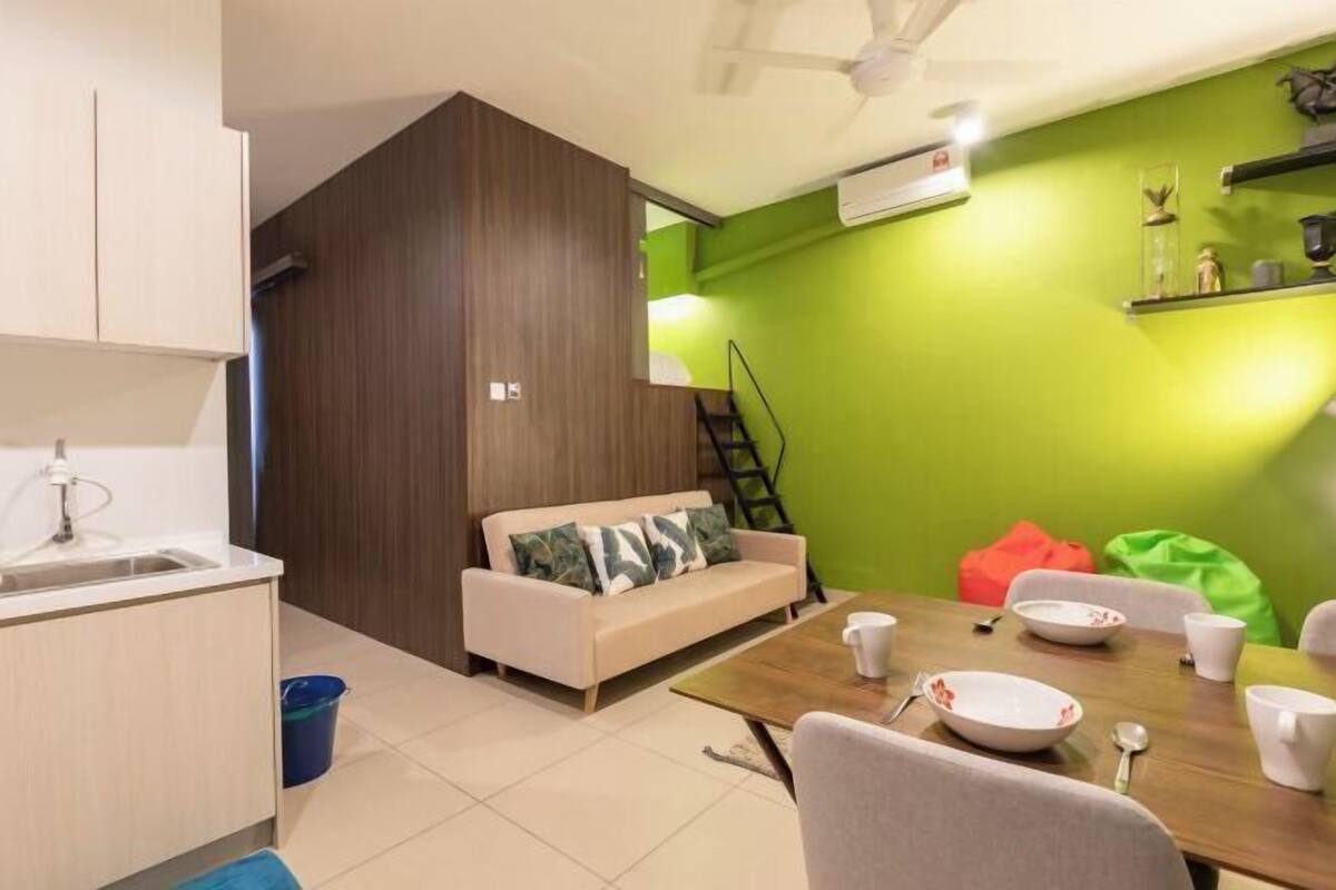 A compact living space with a kitchenette, a sofa with cushions, a dining table with chairs, and a green accent wall.
