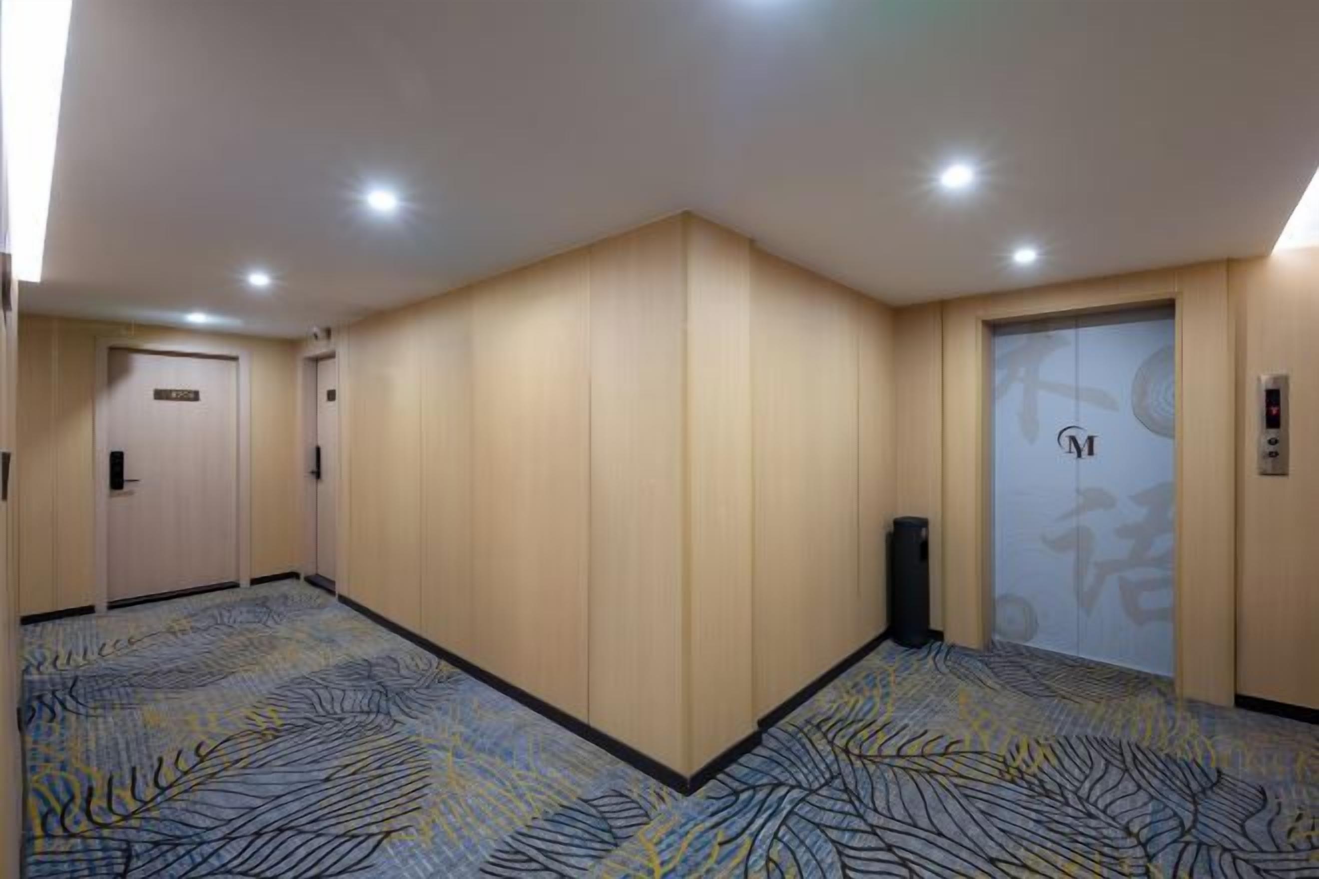 Pasillo de hotel con alfombra estampada, una puerta con placa de identificación y un panel de control en la pared.