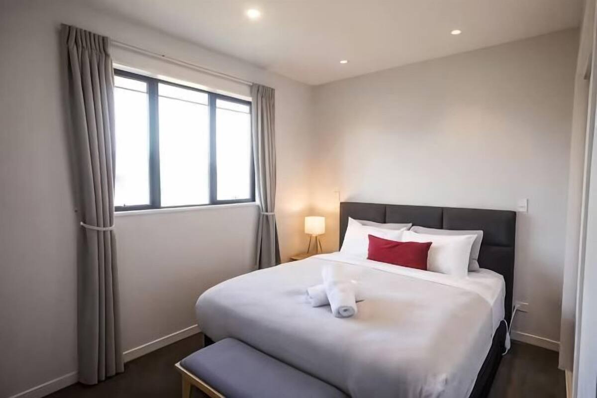 Una habitación de hotel con cama, lámpara de noche, ventana con cortinas y un banco.