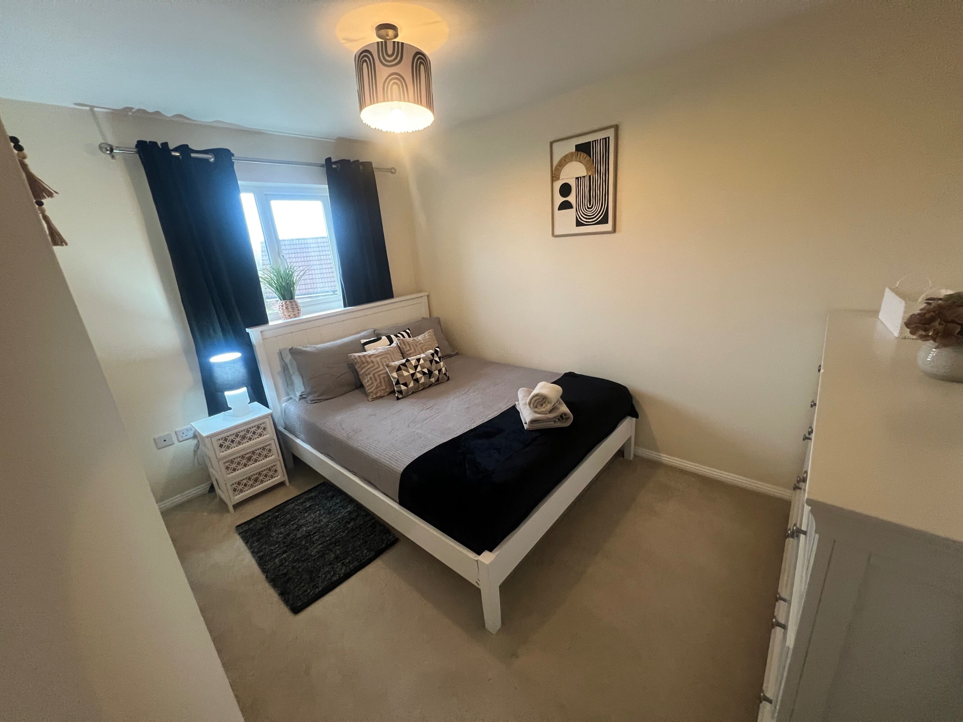 A bedroom with a bed, a nightstand, a dresser, a window with curtains, a wall art piece, and a ceiling light fixture.