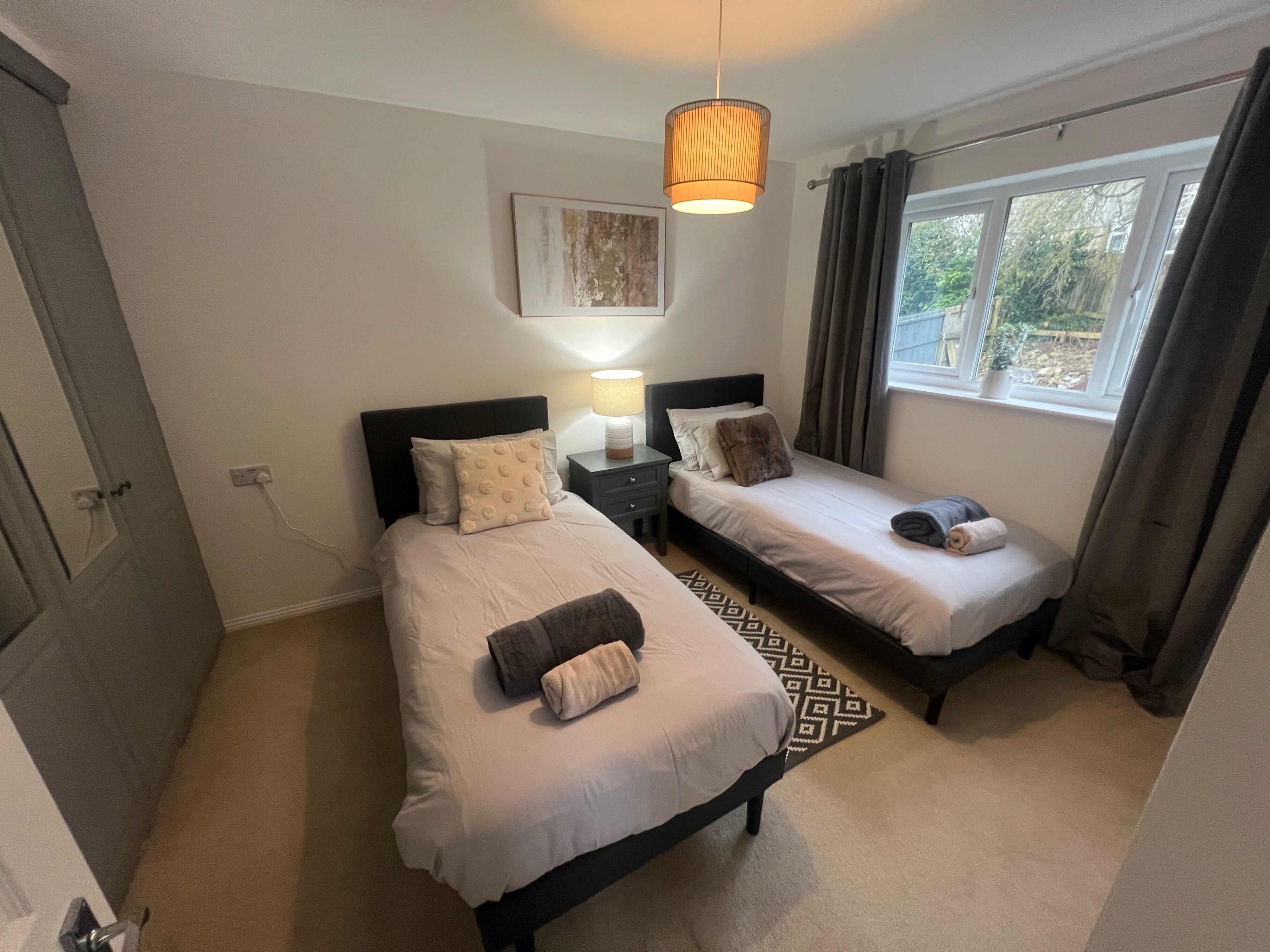 Habitación de hotel con dos camas individuales, una mesita de noche con lámpara, una ventana con cortinas y una lámpara colgante.