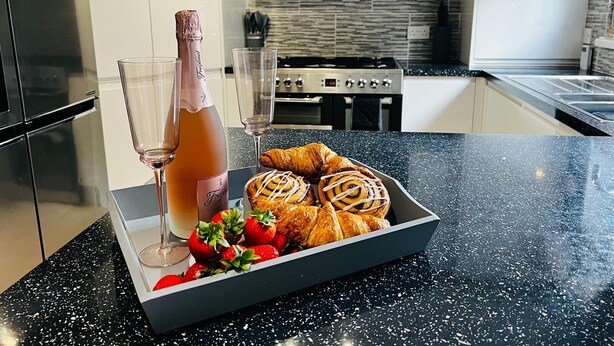 A modern kitchen with a black countertop, a bottle of champagne, glasses, and pastries. - Luxury 4 bed House Sleeps 8, Enjoy in Comfort (Bridgend)
