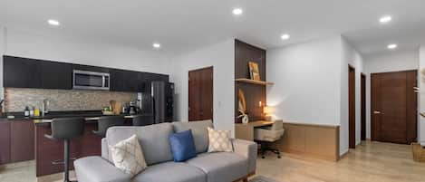 A modern living room with a grey sofa, a coffee table, and a kitchen area with dark cabinets and stainless steel appliances.