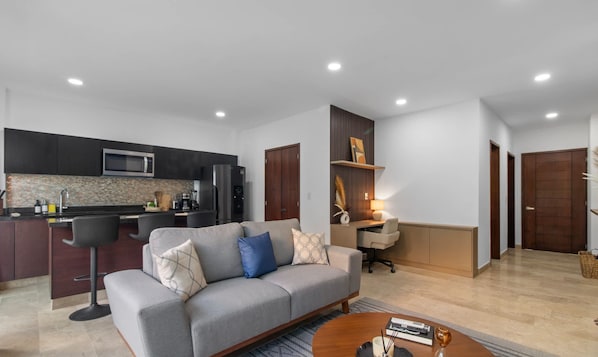 A modern living room with a grey sofa, a coffee table, and a kitchen area with dark cabinets and stainless steel appliances. - Risco - Downtown Bucerias, Rooftop Pool. (Bucerías)