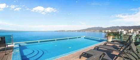 A swimming pool with a view of the sea, lounge chairs, and a wooden deck.