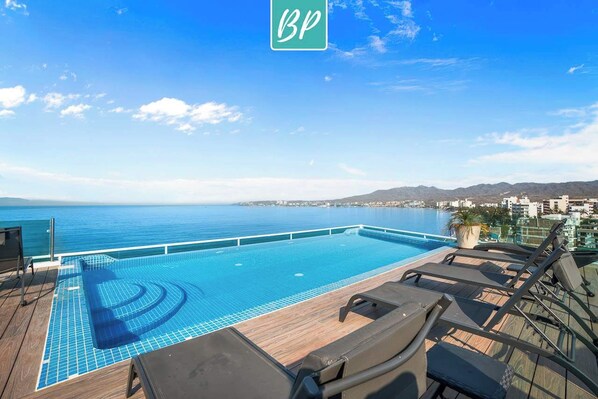 A swimming pool with a view of the sea, lounge chairs, and a wooden deck.