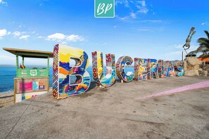 Uma placa colorida com a inscrição 'BUGERMANS' numa praia, com um quiosque e uma palmeira ao fundo.