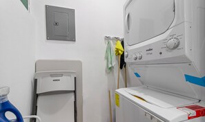 Uma lavanderia com máquina de lavar frontal, secadora e painel elétrico fixado na parede.