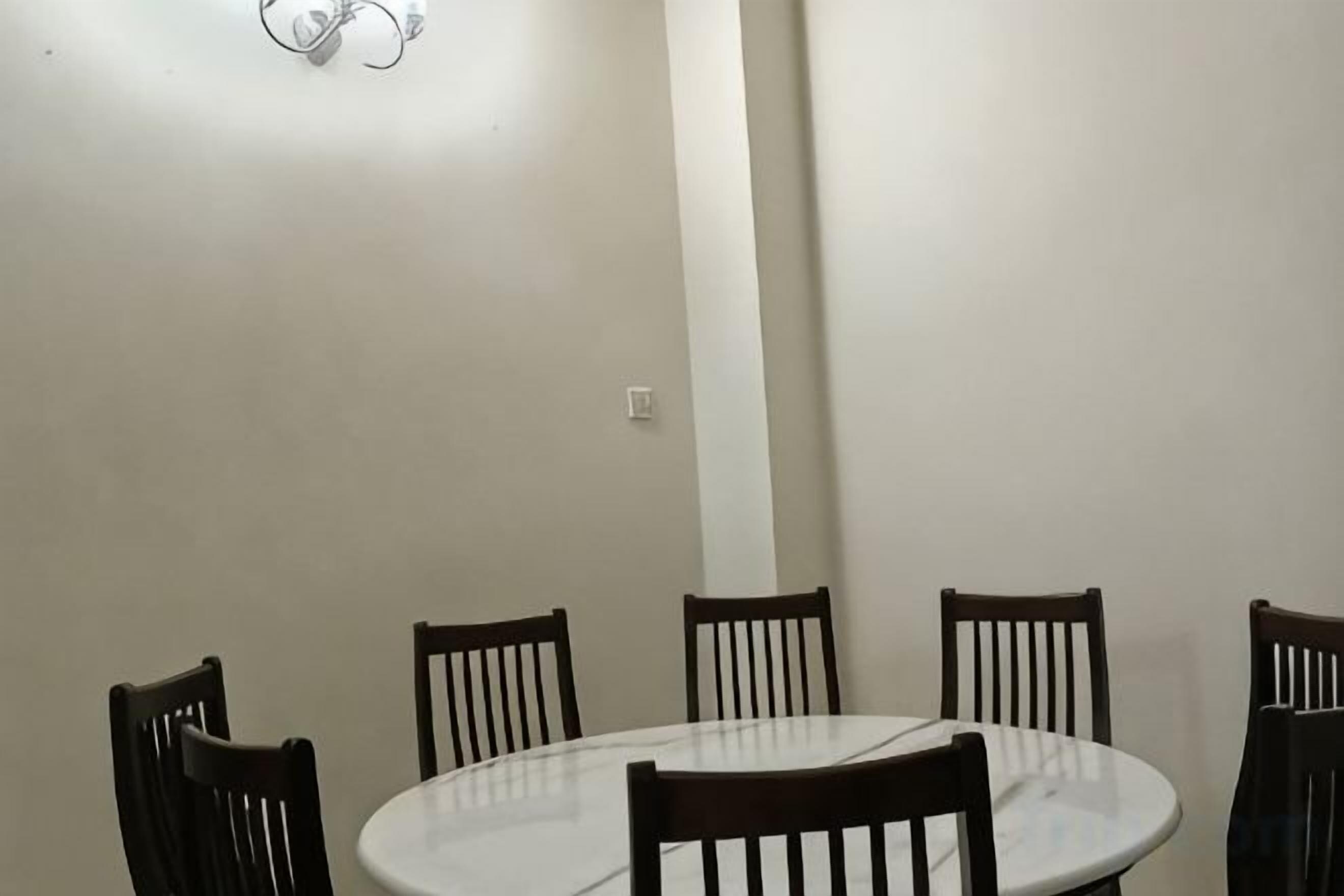 A round dining table with six wooden chairs, a white wall, and a light fixture.