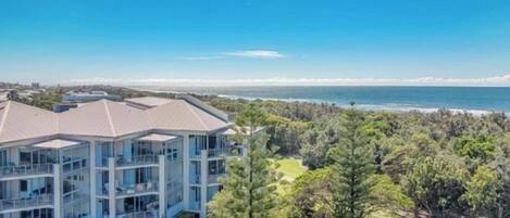 A multi-story building with a flat roof, surrounded by dense greenery and a view of the ocean.