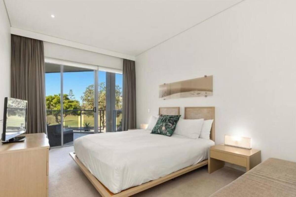 A modern hotel room with a large bed, a bedside table with a lamp, a TV, and a sliding glass door to the balcony.