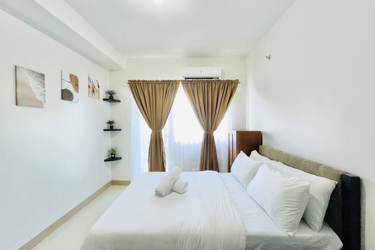 Habitación de hotel con cama, cortinas blancas, ventilador de techo y estantes en la pared con plantas.