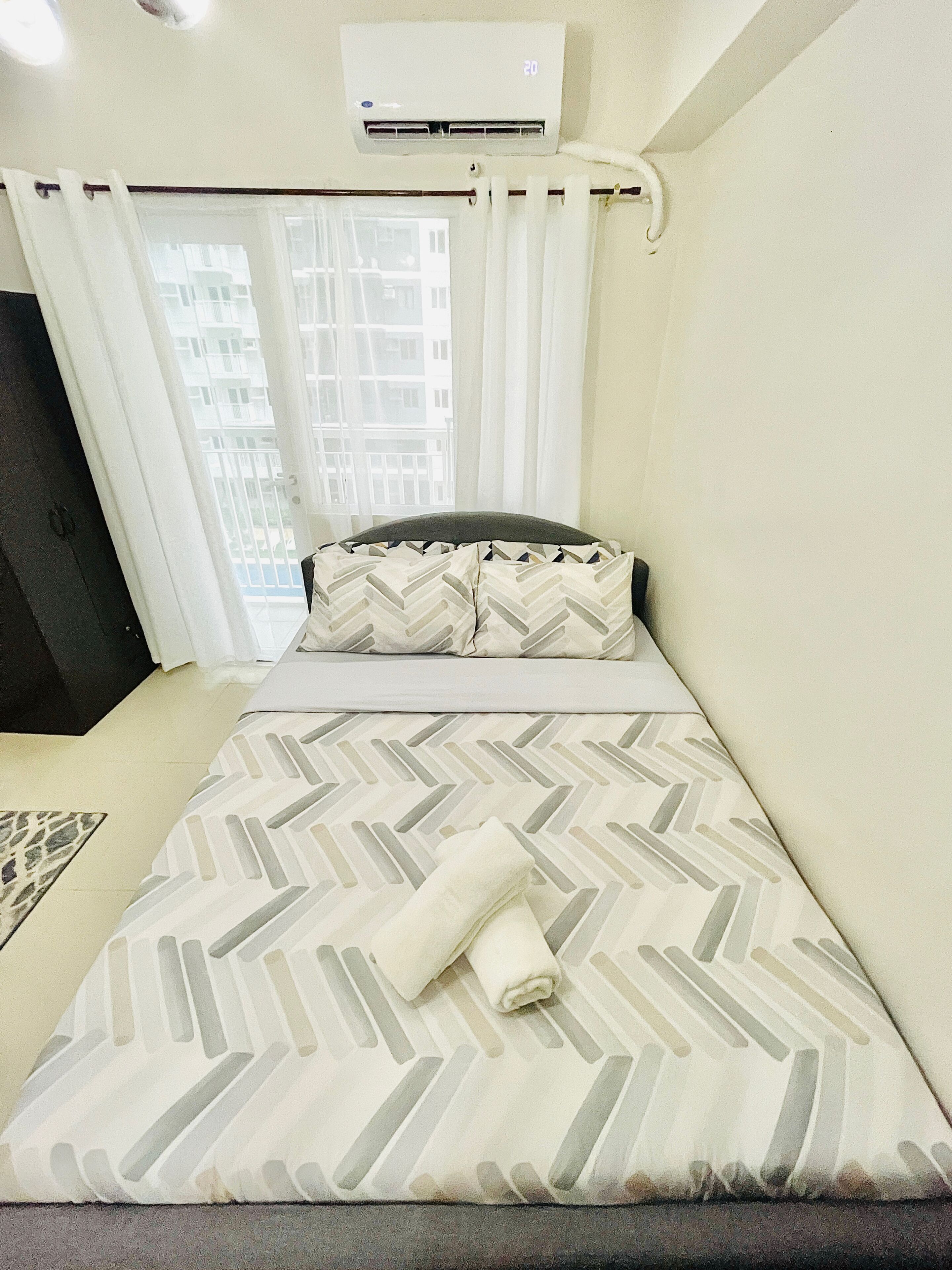 A neatly made bed with a patterned bedspread, a pillow with a chevron design, and a rolled towel. There is a window with curtains, an air conditioning unit above the window, and a view of buildings outside.