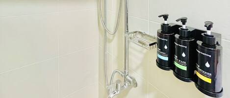 A shower with a handheld showerhead and a wall-mounted faucet. There are three bottles of shampoo and body wash on a shelf.