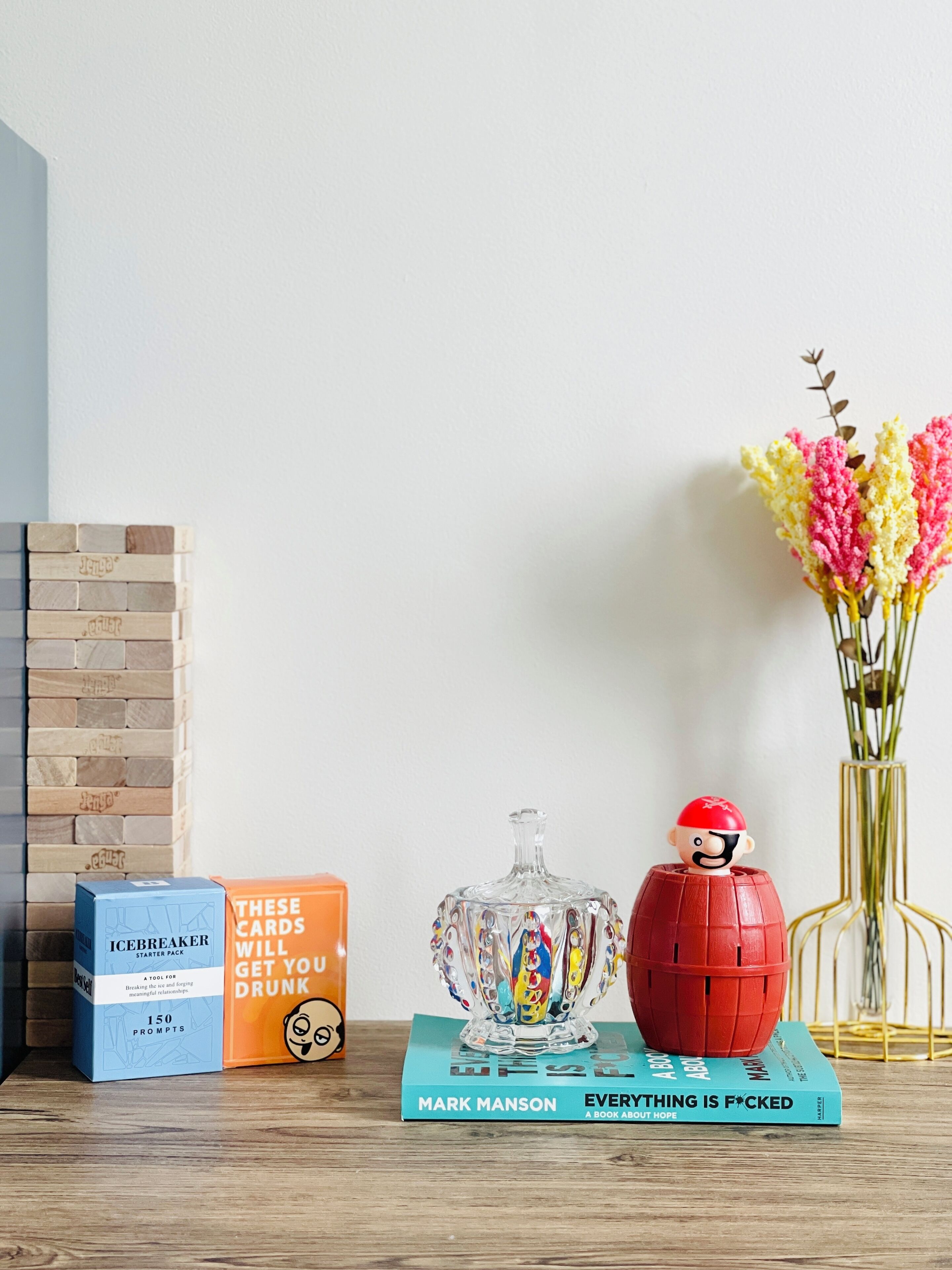 A wooden table with a book titled 'Everything is F*cked', a glass container, a red object, and two boxes labeled 'Icebreaker Starter Pack' and 'These Cards Will Get You Drunk'.