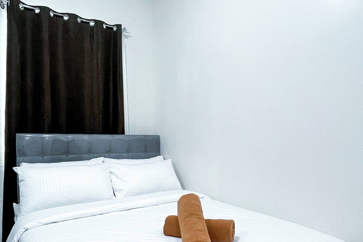 Una cama bien hecha con sábanas blancas y una toalla marrón, una cortina oscura y un cabecero sencillo.